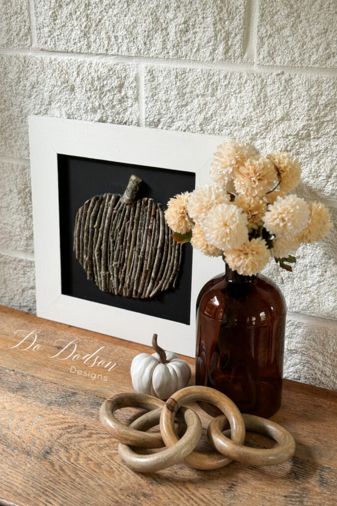 Twig pumpkin decor sitting on a brown wooden table in front of a white brick wall. A brown vase holding white flowers is to the right of the decor, with a white painted pumpkin and a brown wooden chain on the table in front of it